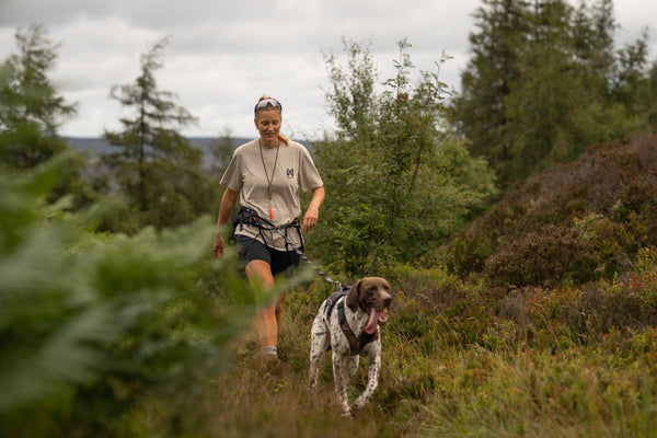 A woman hiking outdoors on a grassy path with a spotted dog, wearing the limited edition t-shirt and dark shorts, surrounded by green trees and shrubs under an overcast sky.